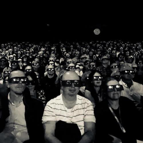 Audience of scary looking people all wearing 3D glasses and looking intently at the stage